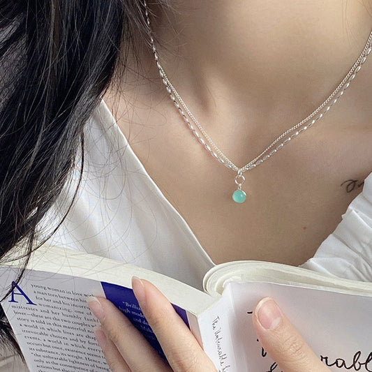 A person wearing a white top and the Aquamarine Natural Stone Necklace Sterling Silver 925, featuring a delicate double-layered design with a small aquamarine pendant, is holding an open book. Only the neck, hand, and part of the book are visible.