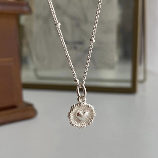 A close-up of the Love Heart Textured Silver 925 Necklace shows its round pendant with a raised heart in the center, hanging against a softly blurred brown and beige background.