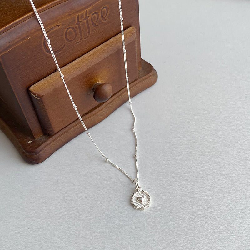 The Love Heart Textured Silver 925 Necklace, featuring a heart-shaped pendant, is displayed on a white surface next to a wooden box labeled Coffee.