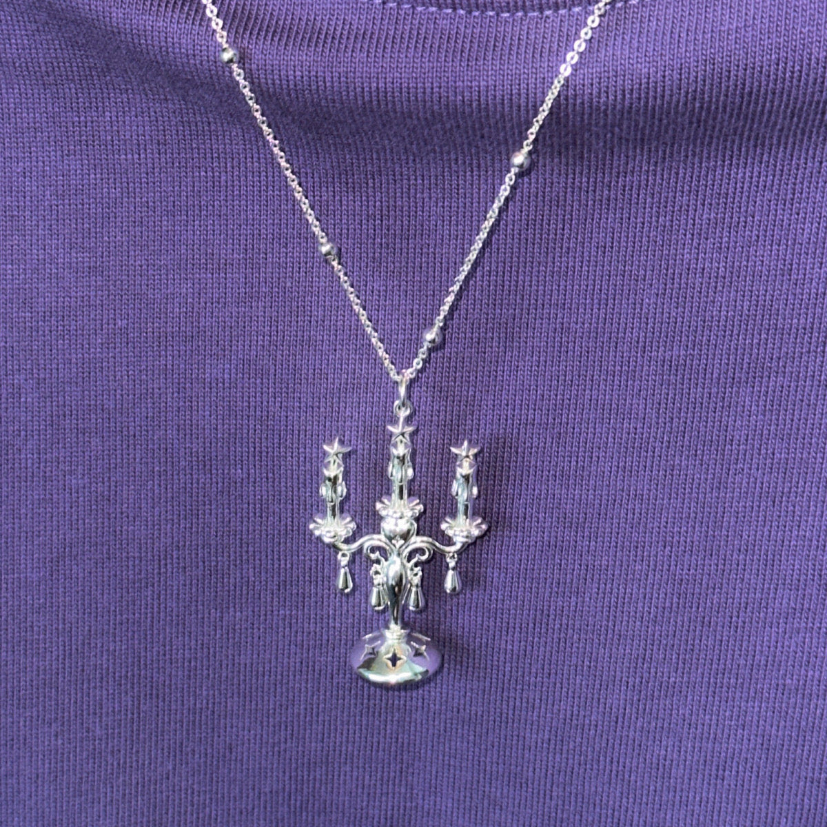 The Trendy Candles decor Style Necklace in Sterling Silver 925 is displayed against a purple ribbed fabric background, featuring a pendant inspired by candle chandelier design.