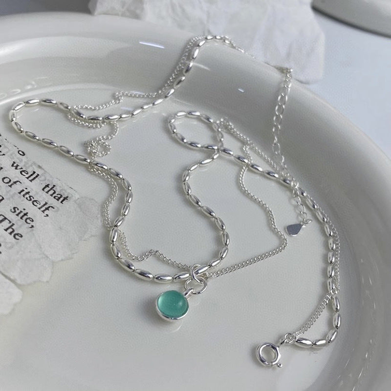 Two sterling silver necklaces rest on a white ceramic dish: one features an aquamarine natural stone pendant, while the other has a delicate chain with a small heart charm. A piece of torn paper with text appears nearby.