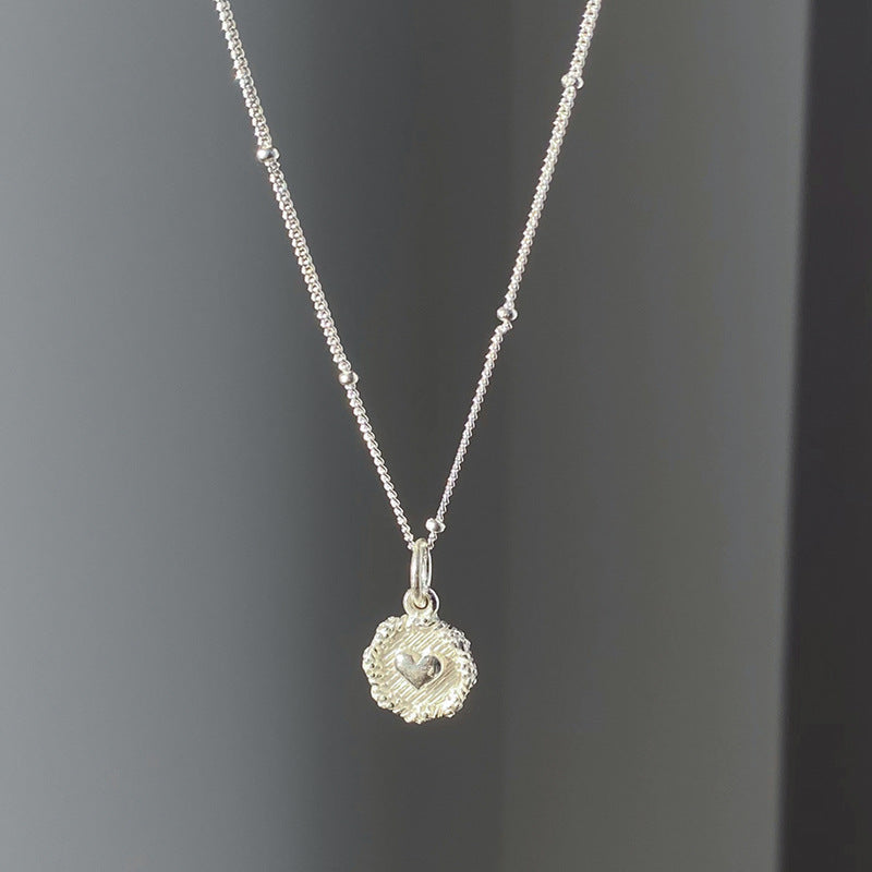 The Love Heart Textured Silver 925 Necklace features a delicate sterling silver chain with a textured round pendant and a small heart design at the center, showcased on a plain dark background.