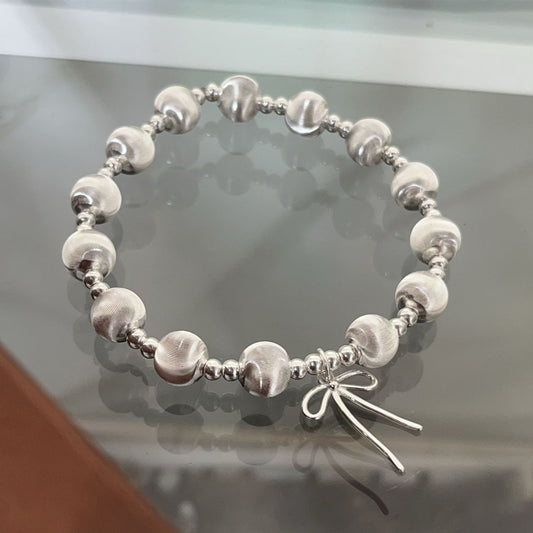 The Heavy weight Beaded 925 Silver Bracelet features alternating smooth and textured round silver beads with a small bow-shaped charm, shown on a reflective glass surface.