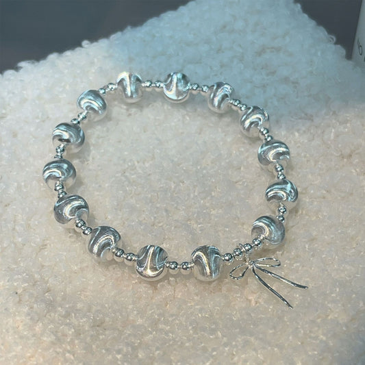 The Heavy Weight Beaded 925 Silver Bracelet features textured, wavy silver beads and a small bow charm, elegantly displayed on a white, textured surface.